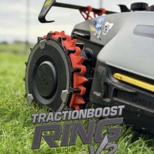 Op de afbeelding: Close-up van een zwart en grijs robotmaaierwiel met een rode tractiering. De tekst "TRACTIONBOOST RING V2" wordt weergegeven in grijs en wit. De robotmaaier staat op groen gras.