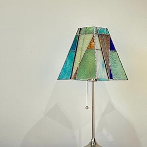 May include: A table lamp with a geometric stained glass shade. The shade features a mosaic of blue, green, and brown glass panels. The lamp has a brushed metal base and a pull chain for the light.