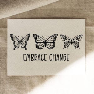 Puede incluir: Una tarjeta color crema presenta tres ilustraciones de mariposas negras sobre la frase "EMBRACE CHANGE" en una fuente manuscrita. Las mariposas tienen diferentes dise&ntilde;os, cada una con patrones de alas detallados.