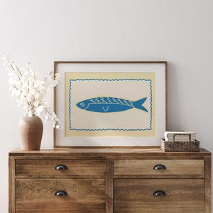 May include: A framed print featuring a blue fish illustration on a cream background, set within a decorative border. The artwork is displayed on a wooden dresser, alongside a vase of white flowers and a small woven basket.