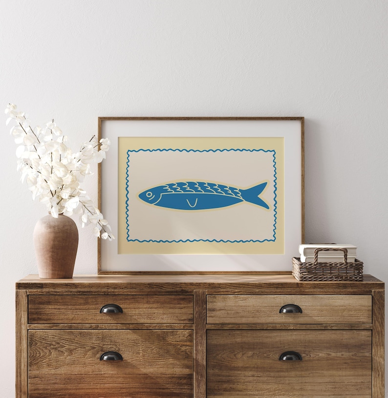 May include: A framed print featuring a blue fish illustration on a cream background, set within a decorative border. The artwork is displayed on a wooden dresser, alongside a vase of white flowers and a small woven basket.
