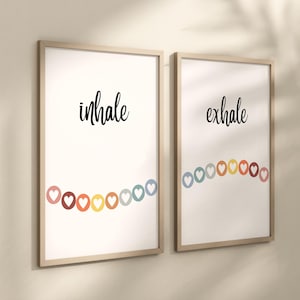 Puede incluir: Dos impresiones art&iacute;sticas enmarcadas con las palabras "inhale" y "exhale" en escritura negra. Cada impresi&oacute;n presenta una cadena de corazones coloridos. Los marcos son de madera clara.