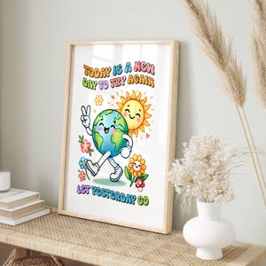 Puede incluir: Una impresi&oacute;n enmarcada con una ilustraci&oacute;n alegre. La obra presenta un personaje de la Tierra sonriente haciendo el signo de la paz, un sol sonriente y el texto "TODAY IS A NEW DAY TO TRY AGAIN LET YESTERDAY GO" en letras coloridas. El marco es marr&oacute;n claro.