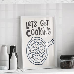 May include: A framed print with the text "LET'S GET COOKING" in black, above a blue line drawing of a frying pan with three fried eggs. The print is displayed on a white countertop with kitchen accessories.