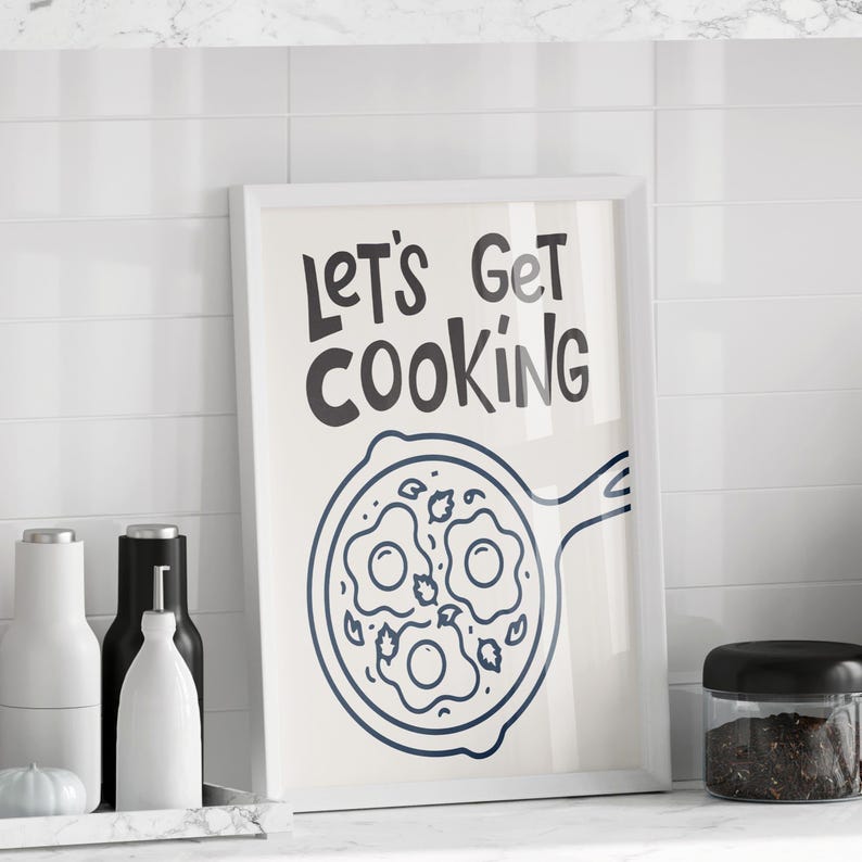 May include: A framed print with the text "LET'S GET COOKING" in black, above a blue line drawing of a frying pan with three fried eggs. The print is displayed on a white countertop with kitchen accessories.