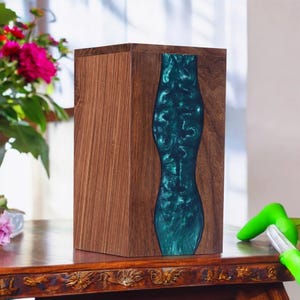 May include: A wooden box with a dark teal resin inlay. The box is made of light brown wood with a rectangular shape. The resin inlay has a flowing, organic design. The box is on a wooden table.