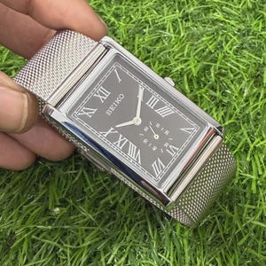 May include: A rectangular Seiko wristwatch with a dark gray dial, silver-tone Roman numeral hour markers, and a silver-tone mesh metal strap. The watch features a small sub-dial for seconds and a polished silver-tone metal case with a side crown.