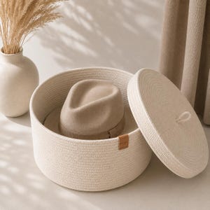 Può includere: Un cesto portaoggetti rotondo in corda intrecciata color bianco sporco con coperchio abbinato e una piccola etichetta in pelle marrone chiaro sul lato. All'interno del cesto si trova un cappello fedora in feltro beige. Design intrecciato e testurizzato.