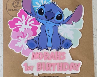 May include: A decorative cake topper featuring a blue Stitch character illustration centered between pink and light blue hibiscus flowers. Below the character, the text reads NORAH'S 1ST BIRTHDAY in pink letters. The topper has two white prongs.