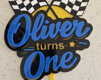 May include: A cake topper featuring two crossed black and white checkered racing flags above the text Oliver turns One in blue and black layered acrylic. The design includes a small yellow star and a yellow stick extending from the bottom.