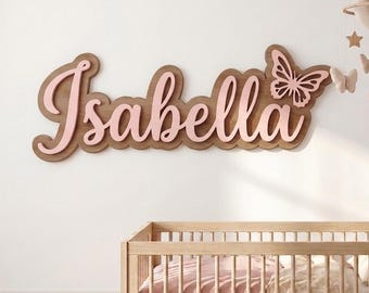 May include: A wooden wall sign featuring the name Isabella in pink script lettering layered over a dark wood backing with a butterfly accent. Above the sign hangs a mobile with a crescent moon, stars, and butterfly shapes in soft pink tones.