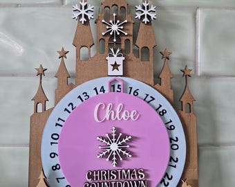 May include: A wooden Christmas countdown calendar featuring a castle silhouette with stars and snowflakes. A central light blue circular dial displays numbers 1 through 24, surrounding a pink circle with the name Chloe, a snowflake, and the text CHRISTMAS COUNTDOWN.