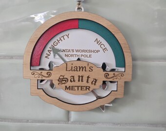May include: A circular wooden hanging ornament featuring a gauge labeled Naughty and Nice with a movable arrow. The center plaque is engraved with the text Liam's Santa METER and SANTA'S WORKSHOP NORTH POLE. It includes a white bead and twine.