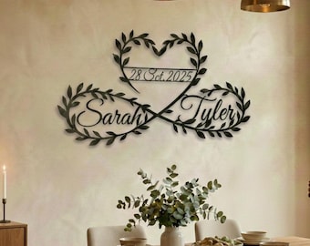 May include: A black metal wall art piece featuring the names "Sarah" and "Tyler" intertwined with an infinity symbol. The date "28 Oct. 2025" is displayed within a heart-shaped design above. The piece is surrounded by leaf-like embellishments.