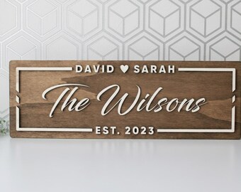May include: A rectangular wooden sign with a dark brown finish and white lettering. The sign reads "David ♥ Sarah" above "The Wilsons" in a cursive font, with "EST. 2023" below. The sign has a white border.