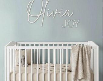 May include: A white crib with a beige mattress and a beige knit blanket draped over the side. Inside the crib sits a small gray plush toy. Mounted on the wall above the crib is a white script sign that reads Olivia JOY in large letters.