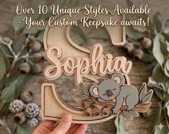 May include: A layered wooden letter S featuring the name Sophia in a light pink script font. A gray koala is depicted sleeping on a branch with green leaves and brown seed pods attached to the letter. Text reads: Over 10 Unique Styles Available - Your Custom Keepsake awaits!