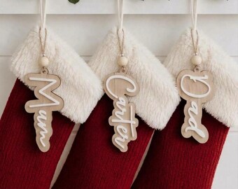 May include: Three red knit Christmas stockings with white faux fur cuffs. Each stocking features a hanging wooden name tag with white script lettering reading Mum, Carter, and Dad, suspended by a light wooden bead and string.