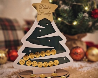 May include: A tree-shaped advent calendar made of dark green and white acrylic with a gold star topper engraved with the text Blake Family. The calendar features slots for gold circular tokens numbered 12 through 25, with several loose tokens scattered at the base.