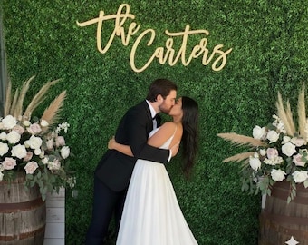 May include: A gold-colored script sign reading "the Carters" mounted on a green artificial boxwood hedge wall. The sign features elegant, flowing cursive lettering and is positioned above a couple kissing in formal wedding attire.