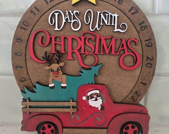 May include: A circular wooden countdown sign featuring the text "DAYS UNTIL CHRISTMAS" with numbers 1 through 25 around the edge. A yellow star sits at the top. A red truck carries a green tree with a reindeer, and Santa is in the driver seat.