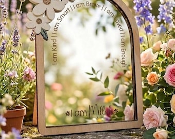 May include: Wooden framed mirror with an arched top, adorned with three white flower cutouts. The mirror's surface reflects a garden scene, and the frame features the phrases "I am Kind," "I am Smart," "I am Brave," "I am Loved," and "I am Enough."