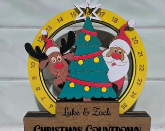 May include: A wooden Christmas countdown calendar featuring a yellow circular ring with numbers 1 through 25, a green Christmas tree, a reindeer, and Santa Claus. The base is engraved with the names Luke & Zack and the text CHRISTMAS COUNTDOWN.