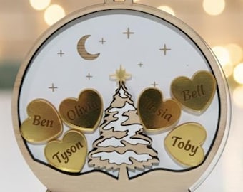 May include: A circular wooden ornament shaped like a snow globe featuring a central Christmas tree, a crescent moon, and stars. Six gold heart-shaped accents are engraved with the names Ben, Olivia, Tyson, Maisie, and Toby. The base reads Hall Family.