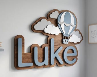 May include: A wall-mounted wooden sign featuring the name Luke in light blue letters layered over a dark wood backing. Above the name is a hot air balloon in blue and white, surrounded by several white and gray cloud-shaped cutouts.