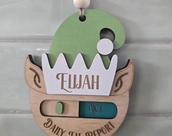 May include: A wooden hanging ornament shaped like an elf head with a green hat and white trim. The name Elijah is engraved on the white brim. Below, a sliding mechanism displays the text Nice in a teal window. The text Daily Elf Report is engraved.