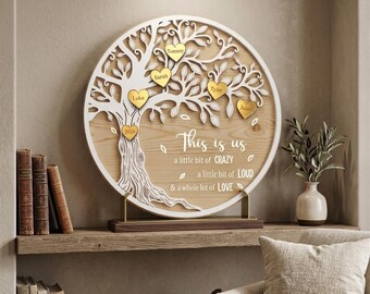 May include: A wooden family tree wall decor with a white tree design and gold heart-shaped name tags. The text "This is us" and other phrases are included. The tree is mounted on a wooden base and displayed on a shelf.