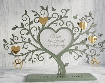 May include: A decorative family tree made of green wood, featuring a heart-shaped photo frame and gold heart-shaped ornaments. The text "Mum, the Heart of our family" is visible. The base reads "OUR FAMILY TREE".