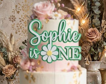 May include: A cake topper featuring the green text "Sophie is ONE" with a white daisy flower replacing the letter "O" in "ONE". The topper has a layered design with a white outline and a green base, positioned on a tiered cake.
