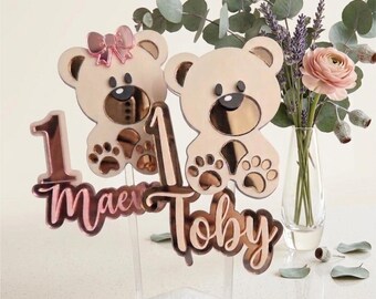 May include: Two custom cake toppers featuring rose gold mirrored acrylic teddy bears. One bear has a pink bow, and both are attached to clear sticks. The toppers display the names Maev and Toby in script font, each accompanied by the number 1.