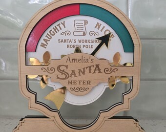 May include: A wooden Santa meter gauge featuring a semi-circular dial with a red Naughty section and a teal Nice section. The center displays the text Amelia's Santa Meter, Santa's Workshop North Pole, and a scroll icon, with a gold arrow pointer.