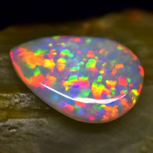 May include: A polished, oval-shaped opal gemstone. The opal displays a milky white base color with vibrant flashes of red, orange, yellow, green, and blue, creating a play-of-color effect. The gemstone is set against a dark background.