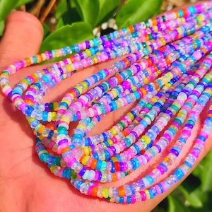 May include: A multi-strand necklace featuring small, circular, flat beads in a vibrant array of colors including neon pink, bright blue, sunny yellow, orange, and translucent white, arranged in a repeating pattern along several thin, flexible cords.