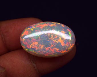 May include: An oval-shaped opal gemstone with a vibrant display of colors, including orange, red, yellow, and green. The gemstone has a polished, glossy surface, and is held in a hand against a dark background.