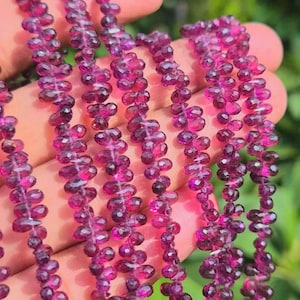 May include: Multiple strands of faceted, deep magenta-colored garnet beads shaped like small teardrops. The beads are strung together in long, vertical lines, showcasing a translucent quality and a reflective, polished surface texture throughout.