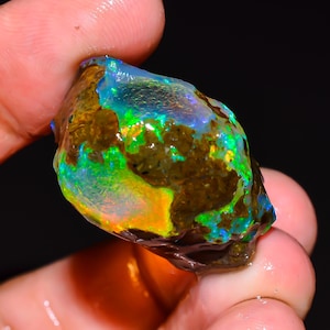 Op de afbeelding: Een ruwe opaal edelsteen met een levendige kleurenpracht. De steen vertoont een mix van groene, blauwe en oranje tinten, met een donkerbruine matrix. De opaal wordt in een hand gehouden, wat de natuurlijke, onbewerkte staat benadrukt.