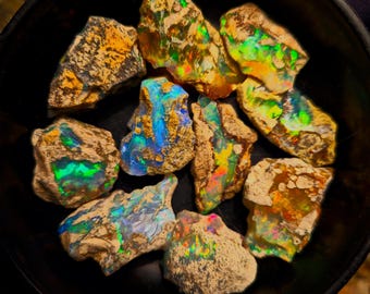 May include: A collection of rough opal gemstones in a black bowl. The opals display vibrant colors including blues, greens, yellows, and oranges. The stones have a natural, textured appearance.