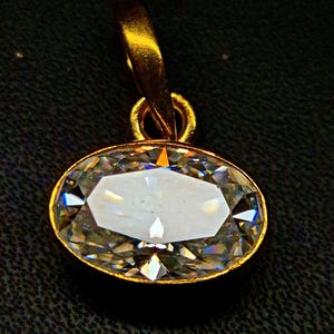 May include: An oval-shaped clear gemstone pendant set in a polished gold-tone metal bezel. The faceted stone displays brilliant light refraction and is attached to a gold-tone metal bail at the top, designed for suspension on a necklace chain.