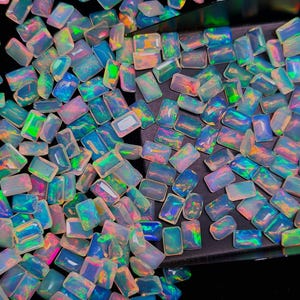Puede incluir: Una colección de piedras preciosas de ópalo iridiscente con cortes rectangulares y ovalados. Los ópalos muestran una gama de colores, incluyendo azules, verdes, rosas y amarillos, con un efecto brillante y multicolor. Las piedras preciosas están dispersas sobre un fondo oscuro.