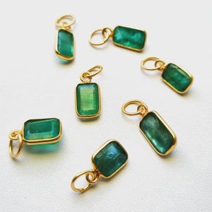 May include: A collection of six rectangular green gemstone pendants, each encased in a gold-toned metal bezel setting with a small circular jump ring attached to the top for hanging. The stones exhibit natural variations in deep green color.