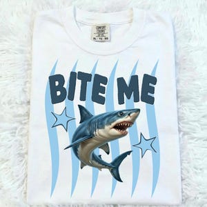 May include: White t-shirt with the text "BITE ME" in dark blue, featuring a detailed illustration of a shark. The design includes blue wavy lines and star shapes. The shirt is folded and appears to be XL-TG-EG size.