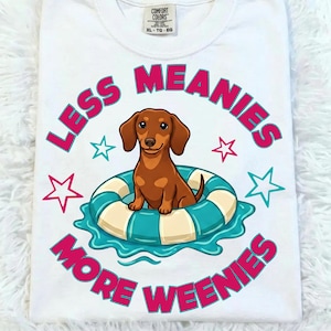 以下が含まれることがあります： 白地のTシャツ。青と白のストライプの浮き輪に乗った、漫画のダックスフンド犬が描かれています。「LESS MEANIES MORE WEENIES」の文字はピンク色で、ピンクとティールの星が周りにあります。