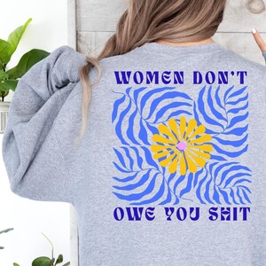 May include: A light gray sweatshirt with the text "WOMEN DON'T OWE YOU SHIT" in purple. The design features a yellow flower with a pink center, set against a blue and white wavy pattern. The sweatshirt is a casual style.