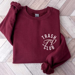 Op de afbeelding: Een bordeauxrode sweatshirt met de tekst "Trash TV Club" in witte, cursieve letters. De sweatshirt is gemaakt van een zacht materiaal en heeft een ronde hals. De mouwen zijn opgevouwen. Het Gildan-merklabel is zichtbaar.