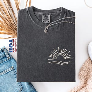 May include: Dark gray t-shirt with the text "Matriarchy Rising" and a sun graphic. The shirt is paired with blue jeans, white sneakers, and a silver necklace. A dried palm leaf and a small USA flag are also in the image.