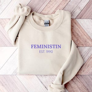 May include: A cream-colored sweatshirt with the word "FEMINISTIN" in purple lettering and the text "EST. 1990" below it. The sweatshirt is folded on a wooden surface.